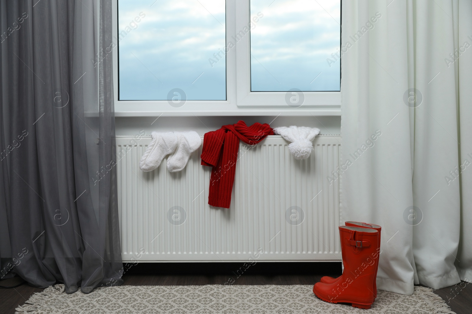 Photo of Heating radiator with hat, socks, scarf and rubber boots near window indoors