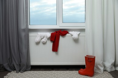 Photo of Heating radiator with hat, socks, scarf and rubber boots near window indoors