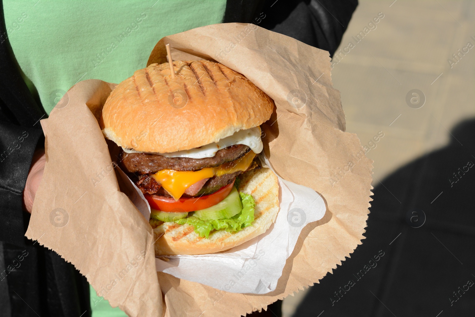 Photo of Woman holding delicious burger in paper wrap outdoors, closeup