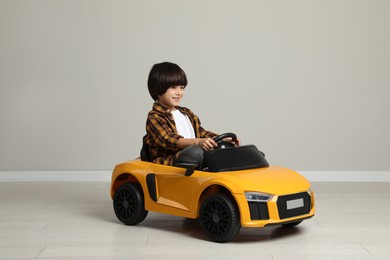 Cute little boy driving children's electric toy car near grey wall indoors