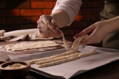 Photo of Woman putting homemade breadsticks on baking sheet at wooden table indoors, closeup. Cooking traditional grissini