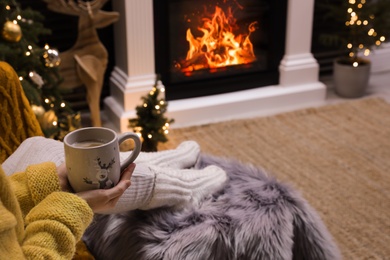 Photo of Woman with cup of drink sitting near burning fireplace at home, closeup