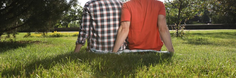 Gay couple sitting on green grass in park, back view. Banner design