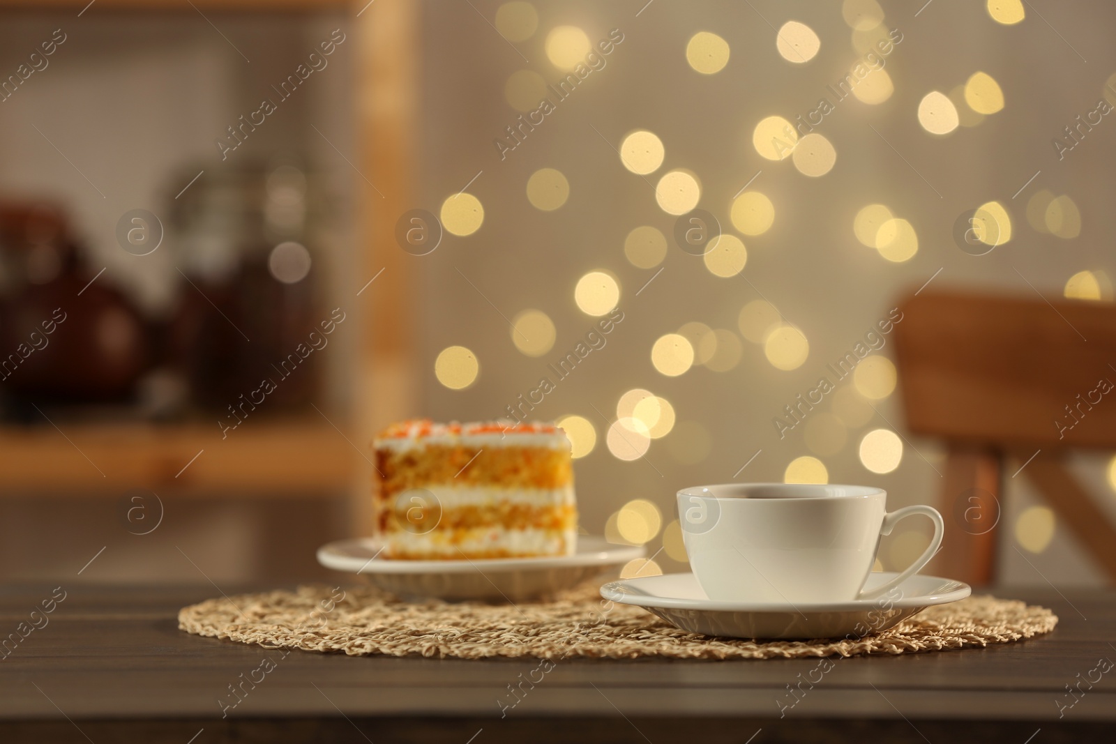 Photo of Hot drink in cup and piece of cake on wooden table against blurred lights indoors