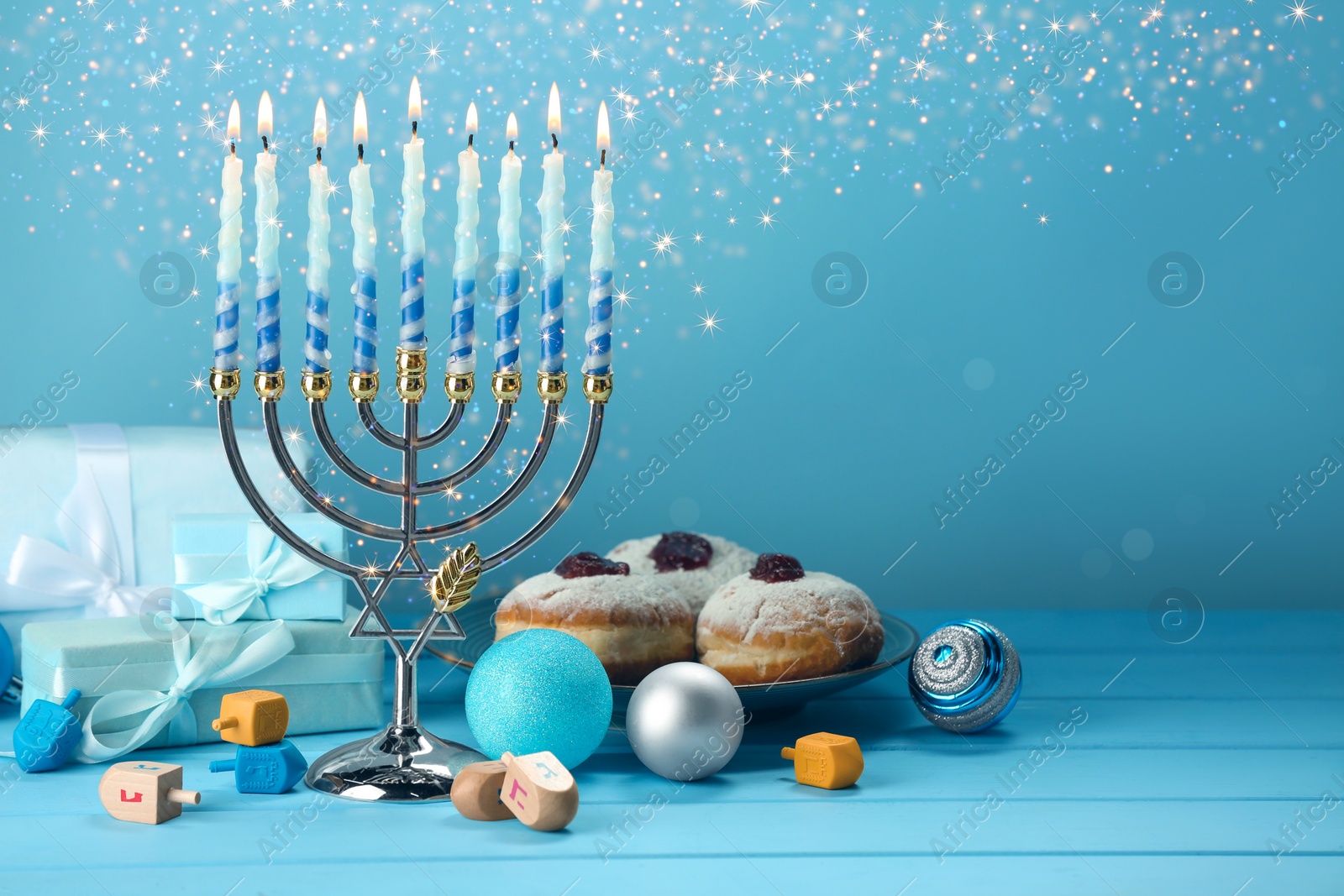 Image of Hanukkah celebration. Menorah with burning candles, dreidels, gift boxes and donuts on light blue wooden table, space for text