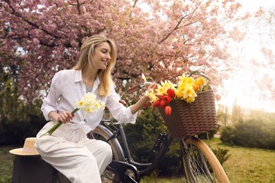 Photo of Beautiful young woman with bicycle and flowers in park on pleasant spring day