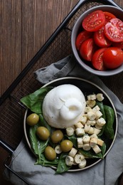 Photo of Delicious burrata cheese served with olives, croutons, basil and tomatoes on wooden table, top view