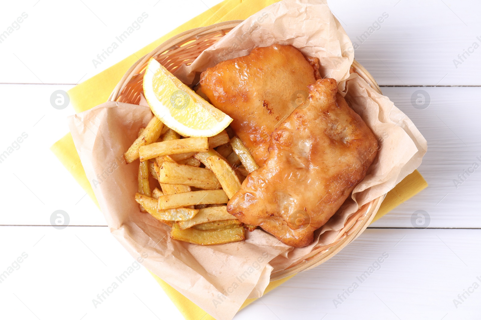 Photo of Tasty fish, chips and lemon in wicker bowl on white wooden table, top view