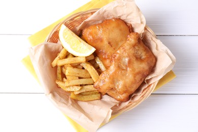 Photo of Tasty fish, chips and lemon in wicker bowl on white wooden table, top view