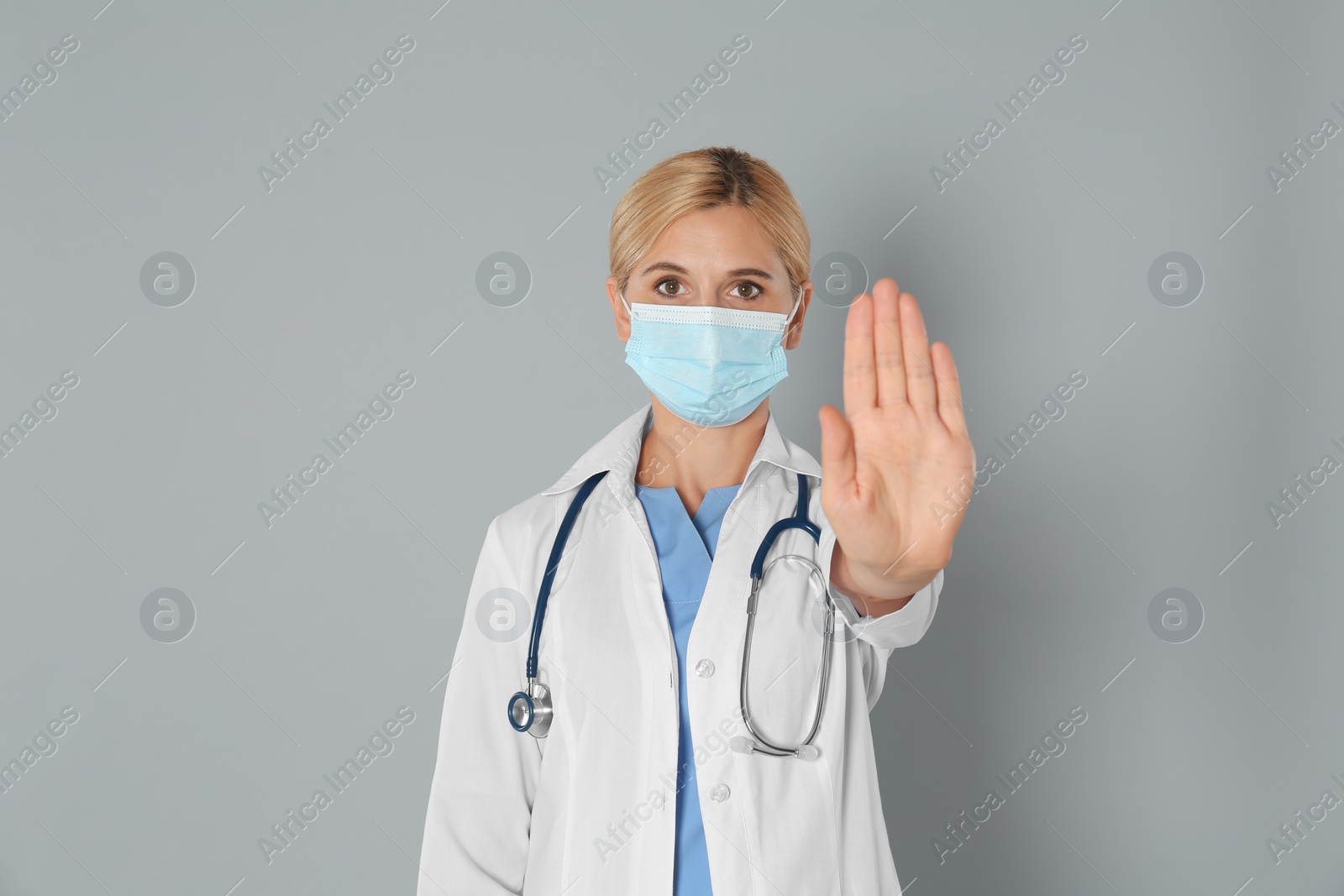 Photo of Doctor in protective mask showing stop gesture on grey background. Prevent spreading of coronavirus