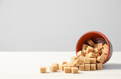 Photo of Overturned bowl with brown sugar cubes on white wooden table, space for text