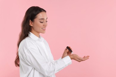 Beautiful happy woman with roller bottle applying essential oil onto wrist on pink background