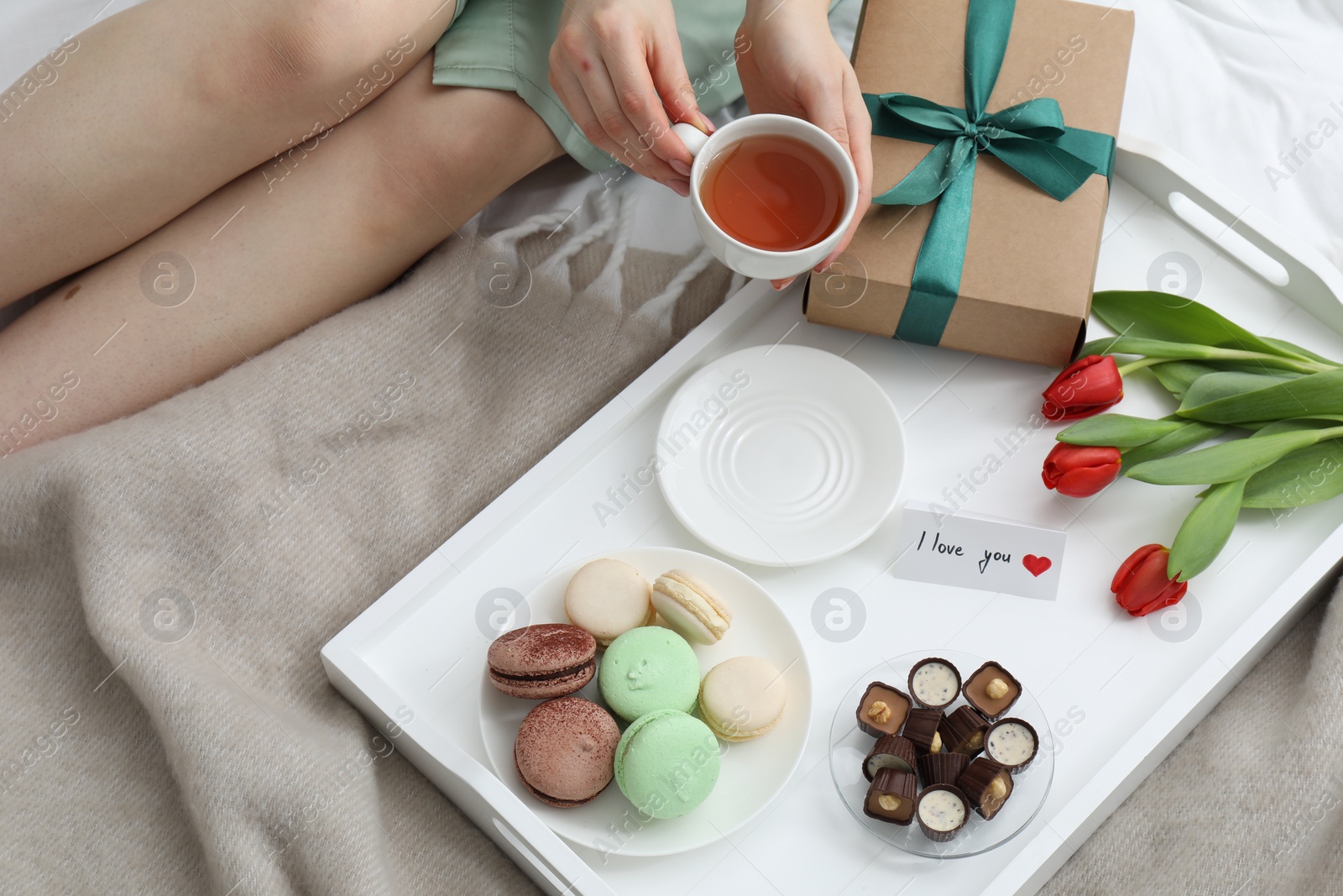 Photo of Tasty breakfast served in bed. Woman with tea, desserts, gift box, flowers and I Love You card at home, closeup