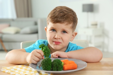 Photo of Displeased little boy eating vegetables at table in room