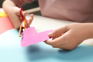 Photo of Child cutting out paper heart with craft scissors at table, closeup