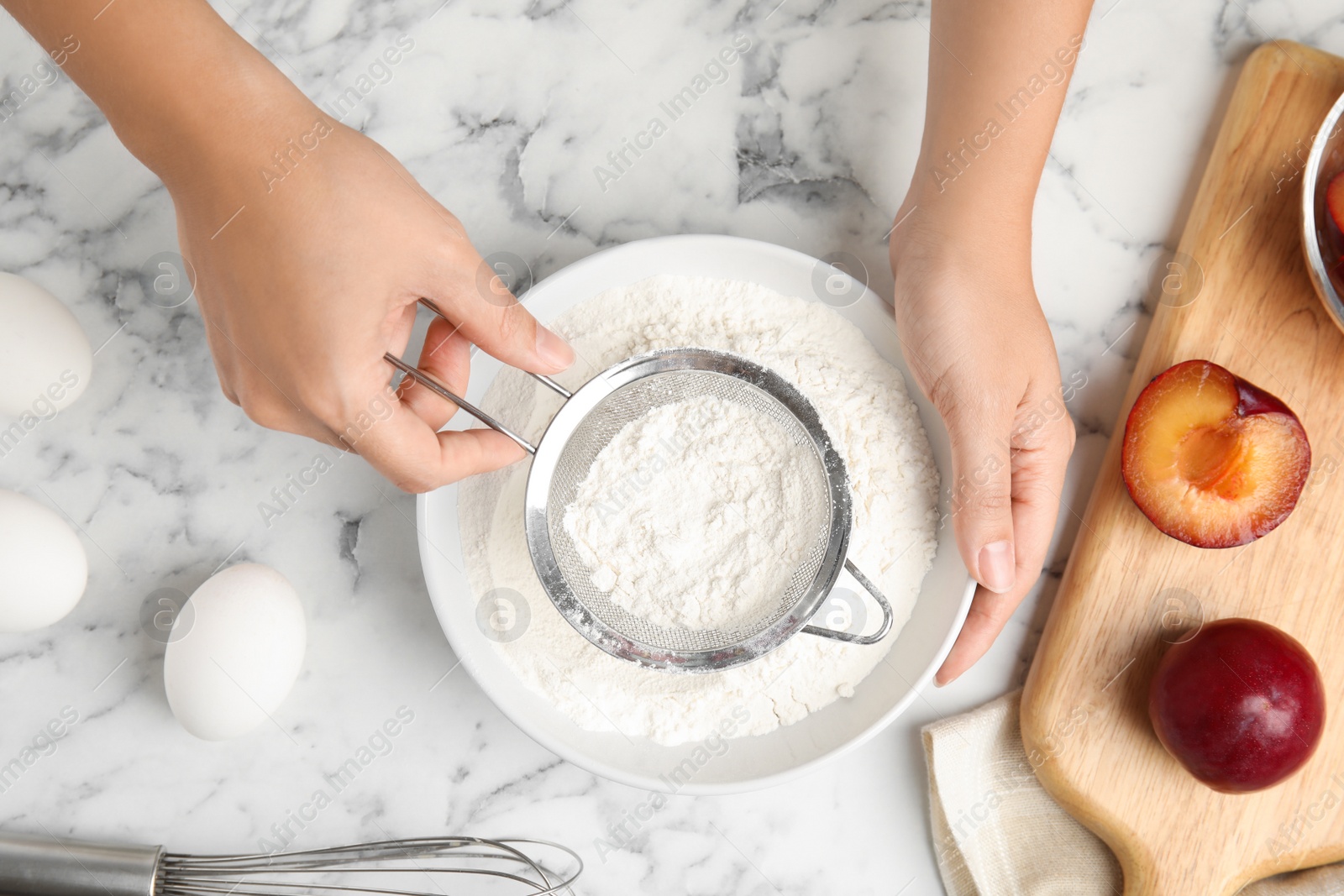 Photo of Woman sieving flour at white marble table, top view. Cooking of delicious plum cake