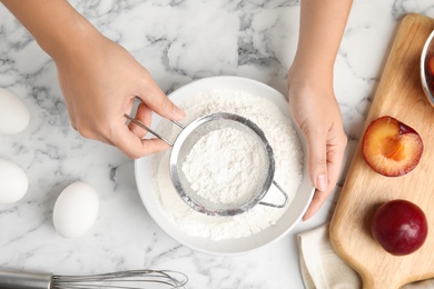 Woman sieving flour at white marble table, top view. Cooking of delicious plum cake