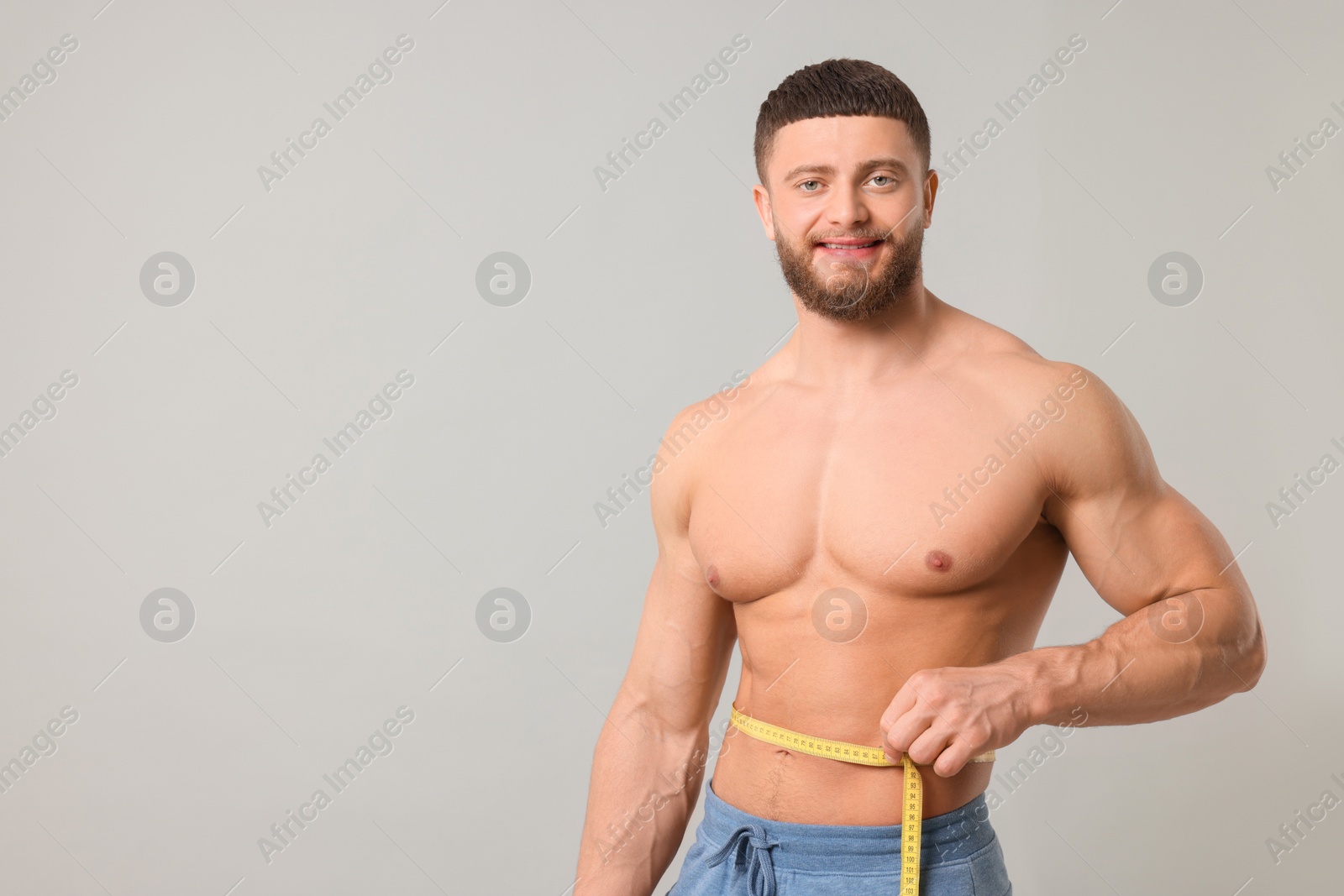 Photo of Portrait of happy athletic man measuring waist with tape on light grey background, space for text. Weight loss concept