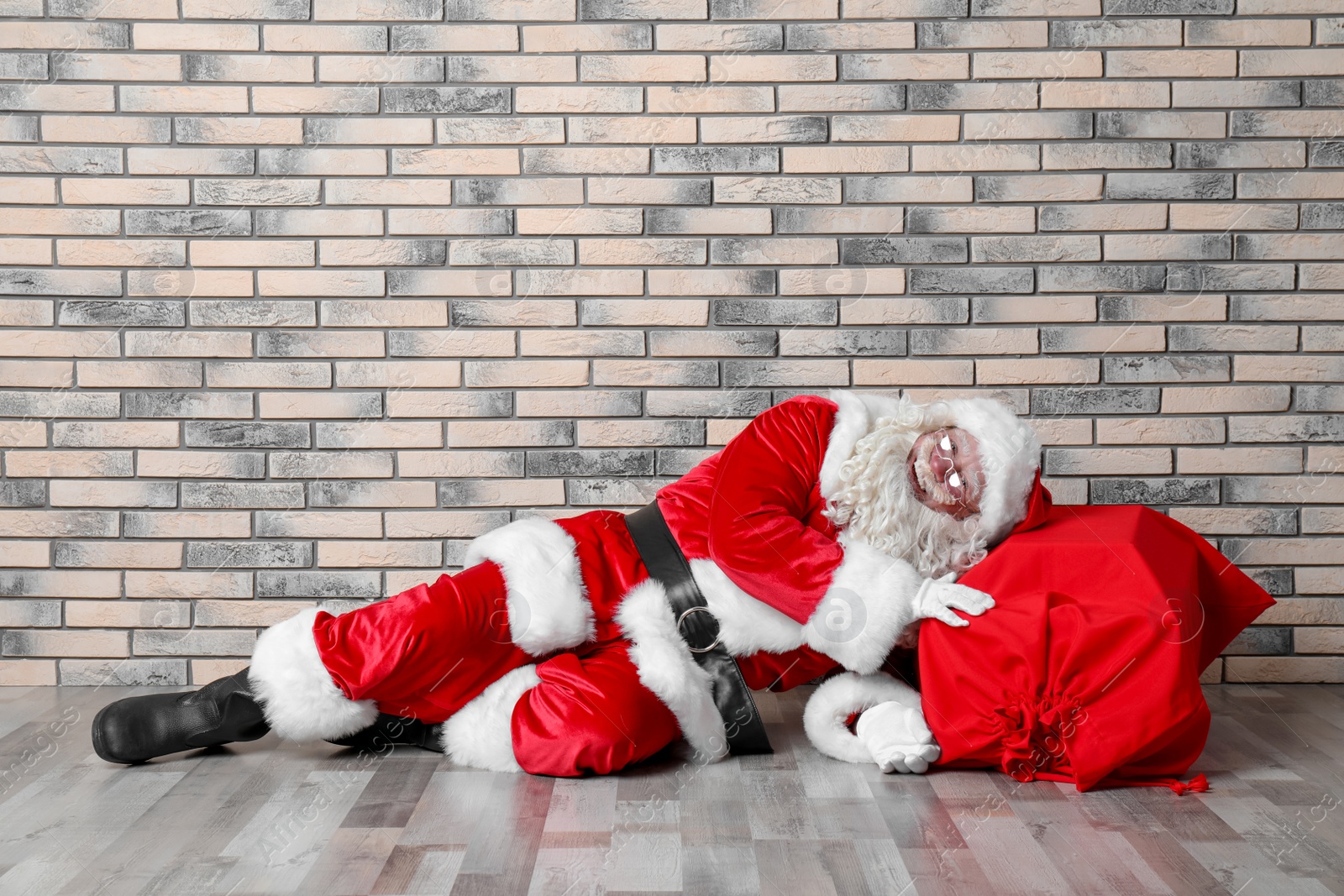 Photo of Authentic Santa Claus with big red bag full of gifts lying on floor near brick wall