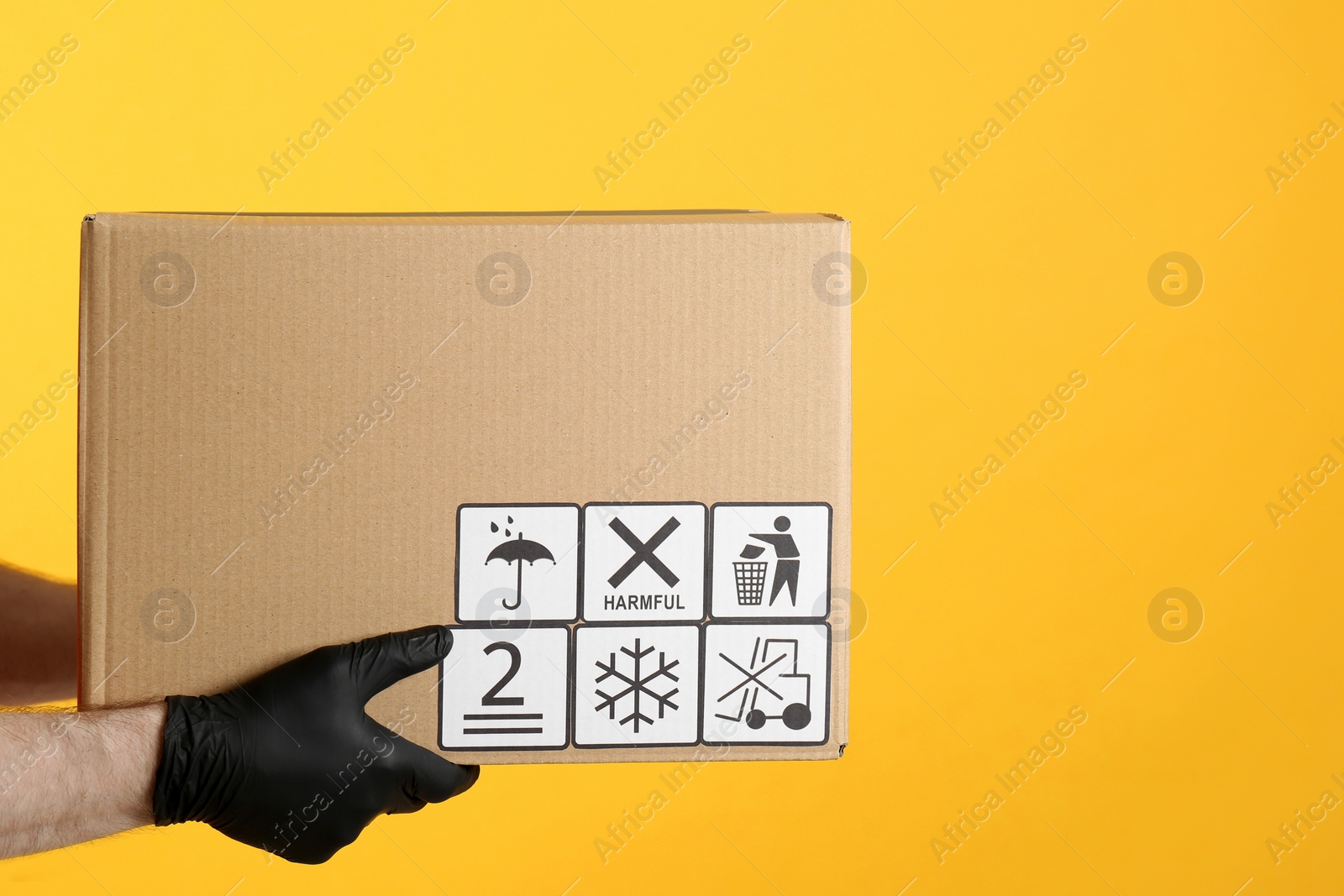 Photo of Courier holding cardboard box with different packaging symbols on yellow background, closeup. Parcel delivery