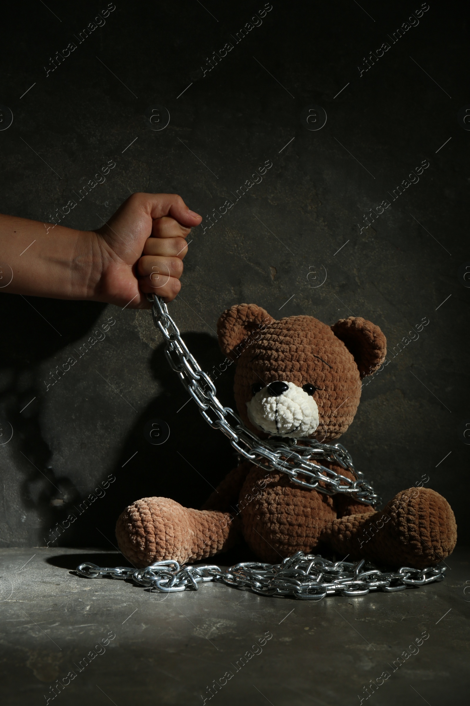 Photo of Stop child abuse. Man holding chain with toy bear in dark room, closeup