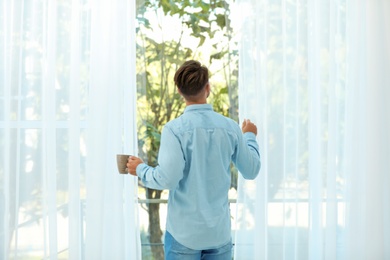 Young man standing near window with beautiful curtains at home