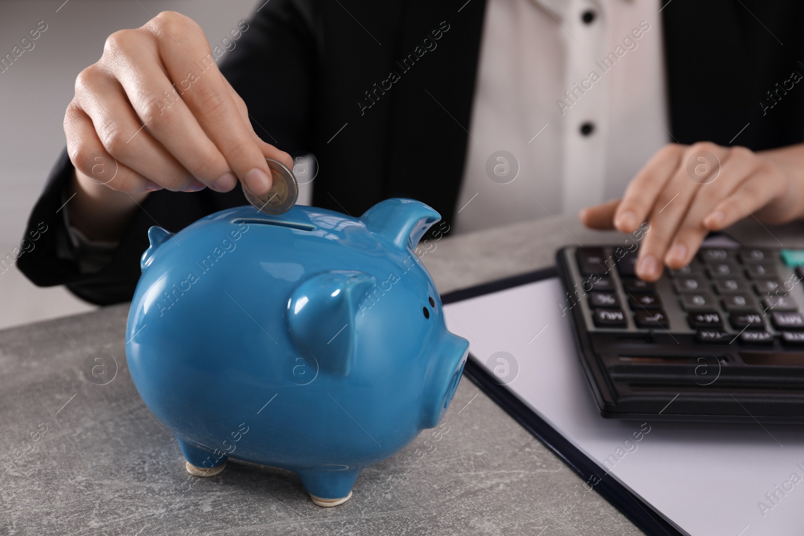 Photo of Woman with calculator putting money into piggy bank at grey table, closeup