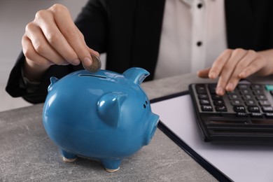 Photo of Woman with calculator putting money into piggy bank at grey table, closeup