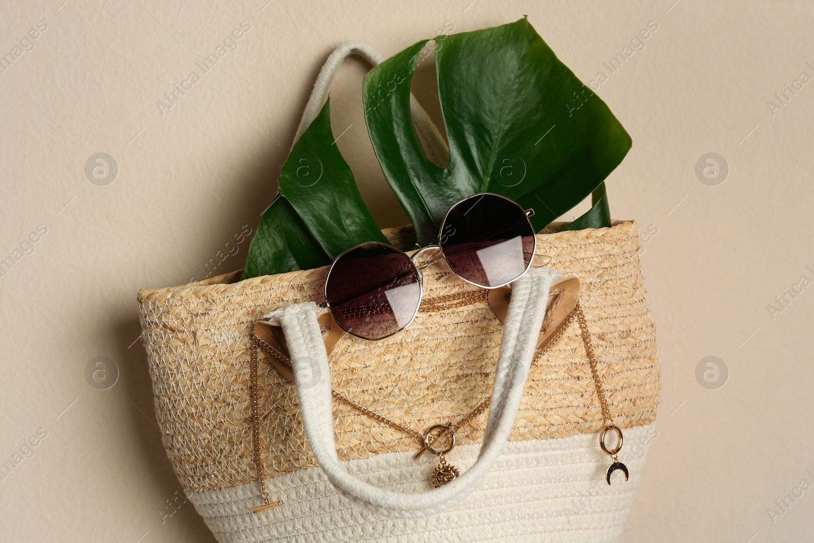 Photo of Elegant woman's straw bag with tropical leaf and accessories hanging on beige background