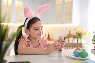 Photo of Cute little girl in bunny ears headband painting Easter eggs at table indoors