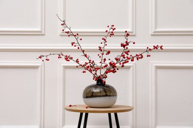 Photo of Hawthorn branches with red berries in vase on wooden table near white wall indoors