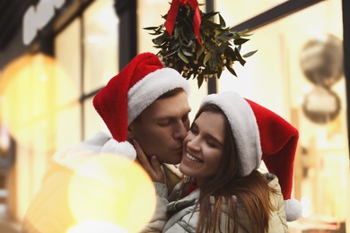 Photo of Happy man kissing his girlfriend under mistletoe bunch outdoors, bokeh effect
