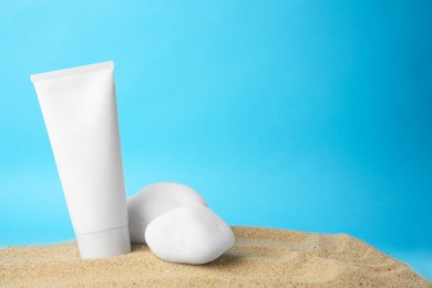 Photo of Tube of cream and stones on sand against light blue background, space for text. Cosmetic product