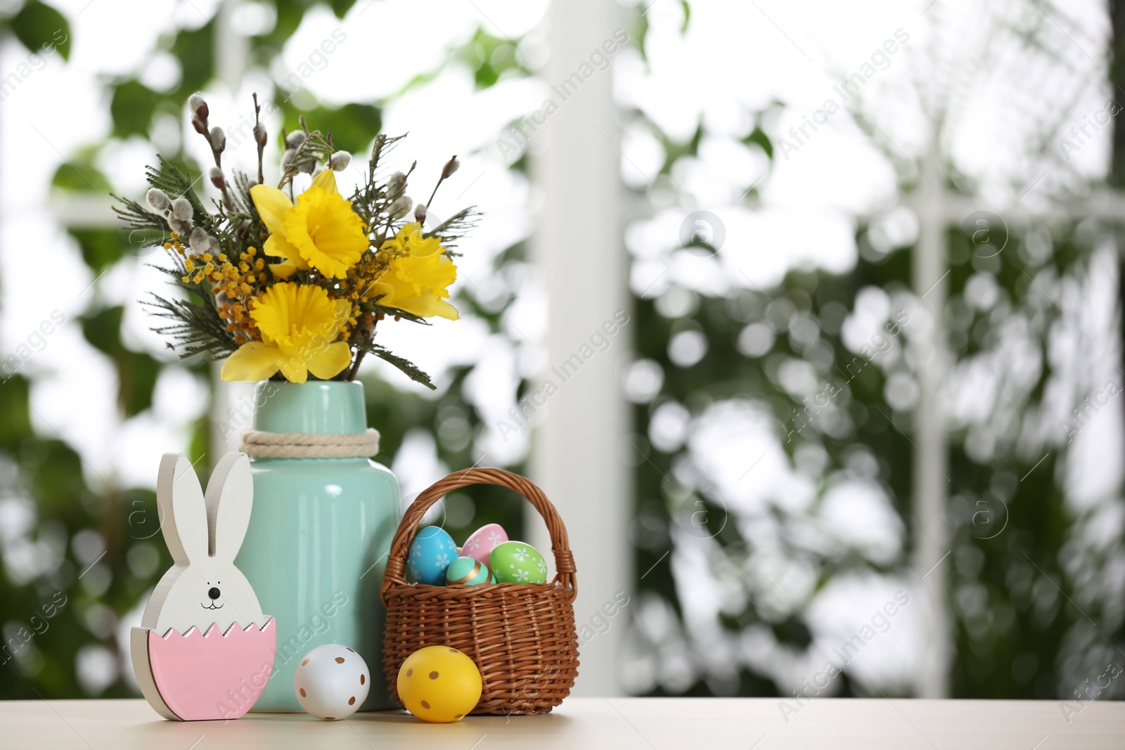 Photo of Festive composition with Easter eggs in wicker basket on table against blurred window, space for text