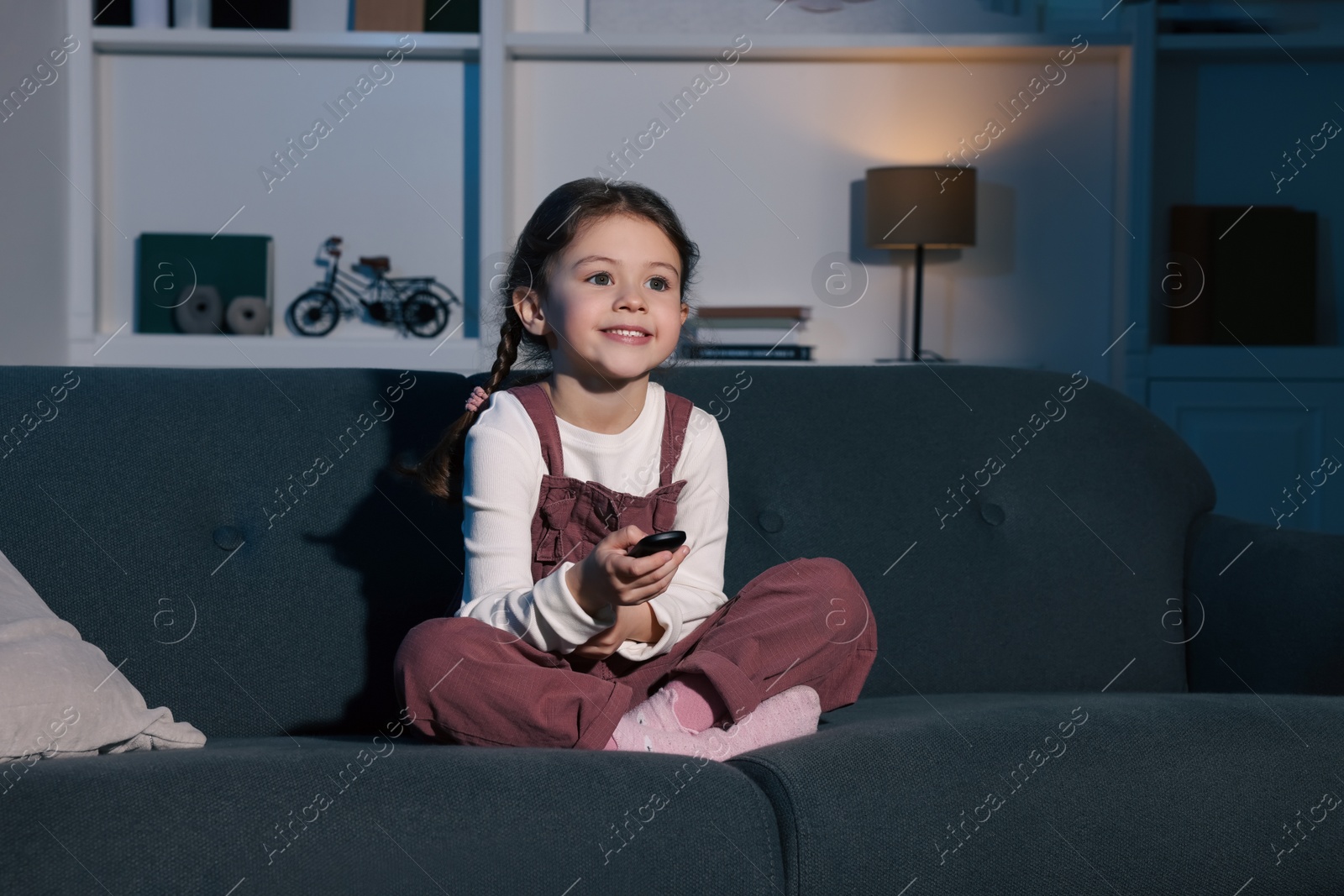 Photo of Happy girl changing TV channels with remote control on sofa at home