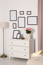 Photo of Empty frames hanging on white wall and chest of drawers with flowers in room