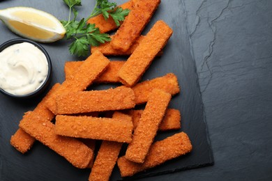 Photo of Fresh breaded fish fingers, sauce and lemon served on black table, top view