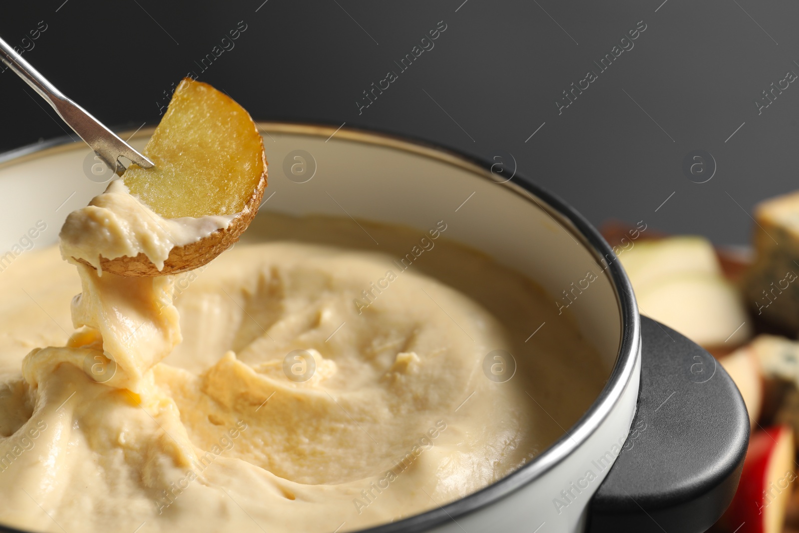 Photo of Dipping piece of potato into fondue pot with melted cheese on grey background, closeup