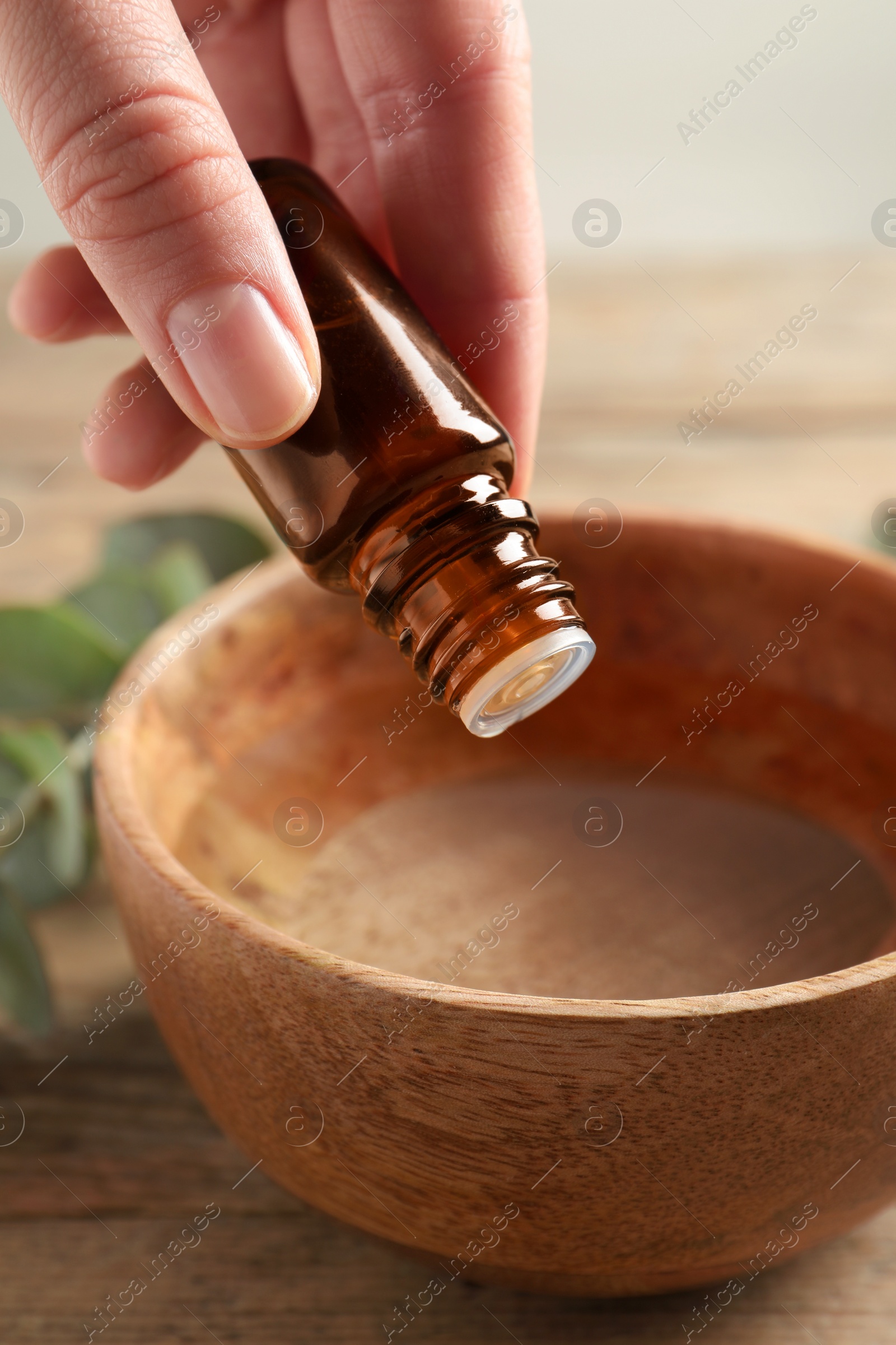 Photo of Woman dripping eucalyptus essential oil from bottle into bowl at wooden table, closeup