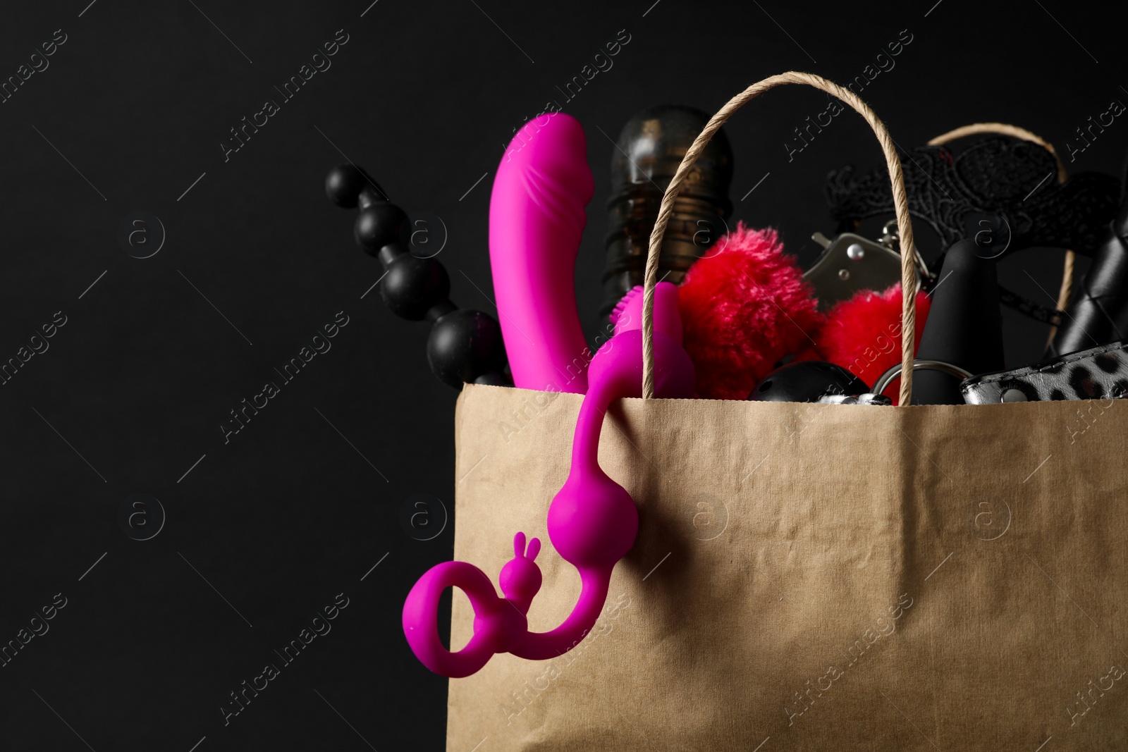 Photo of Paper shopping bag with different sex toys on black background, closeup