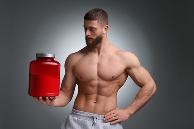 Young man with muscular body holding jar of protein powder on grey background