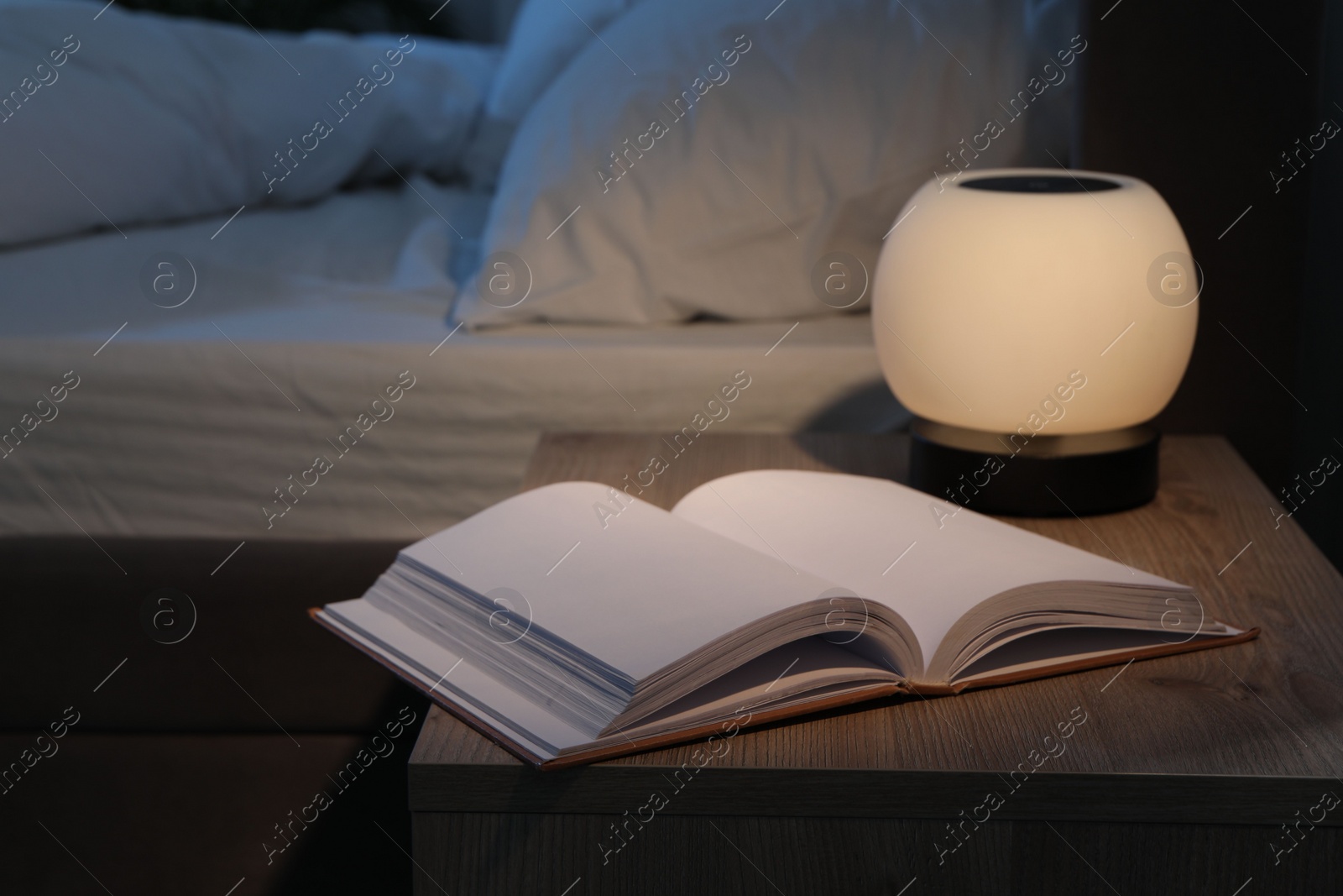 Photo of Stylish nightlight and book on bedside table near bed indoors