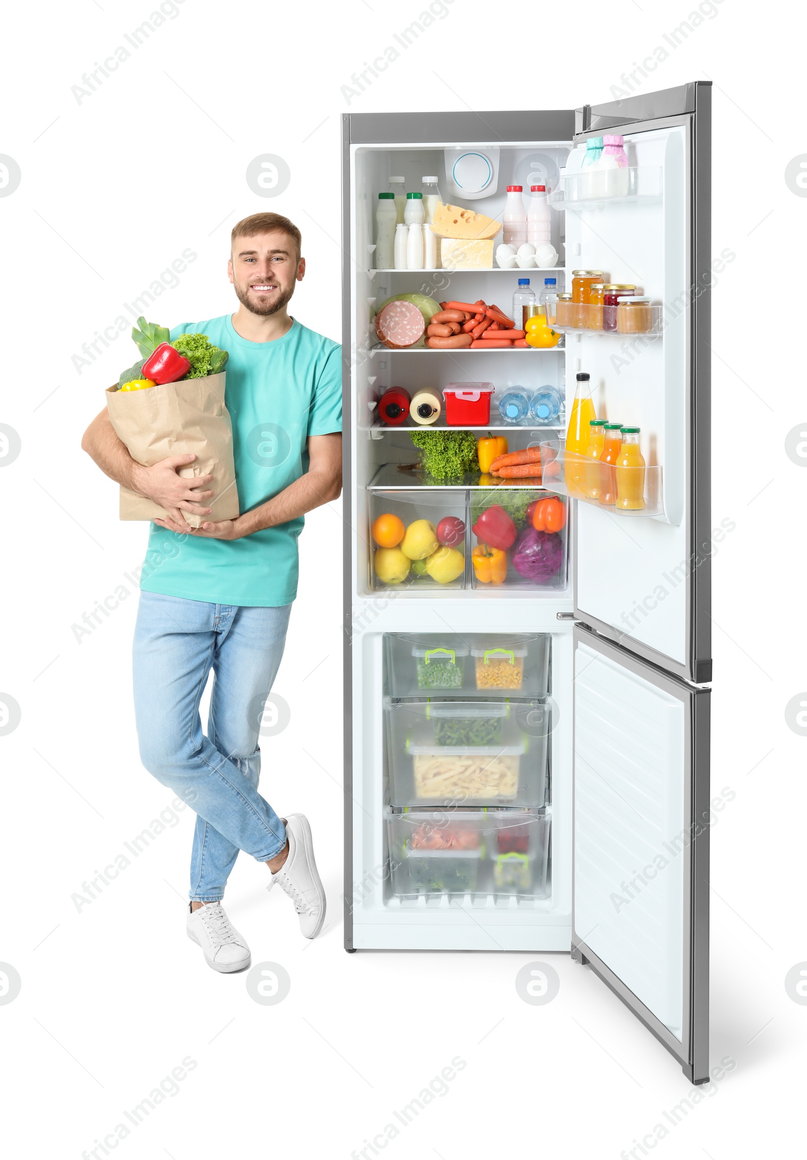 Photo of Young man with bag of groceries near open refrigerator on white background