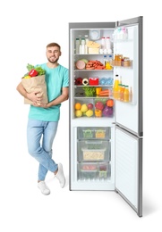 Photo of Young man with bag of groceries near open refrigerator on white background