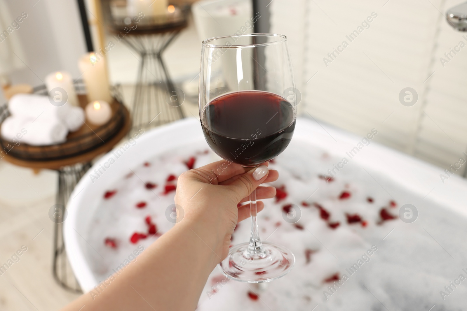 Photo of Woman holding glass of wine against bath tub with foam and rose petals indoors, closeup