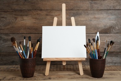 Photo of Easel with blank canvas and brushes on wooden table