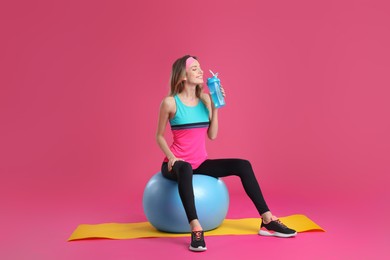 Photo of Beautiful woman sitting on fitness ball and drinking water against pink background