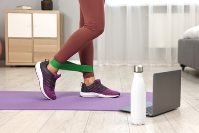 Photo of Online fitness trainer. Woman doing exercise with elastic band near laptop at home, closeup