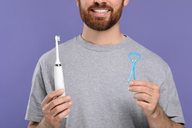 Man with tongue cleaner and electric toothbrush on violet background, closeup