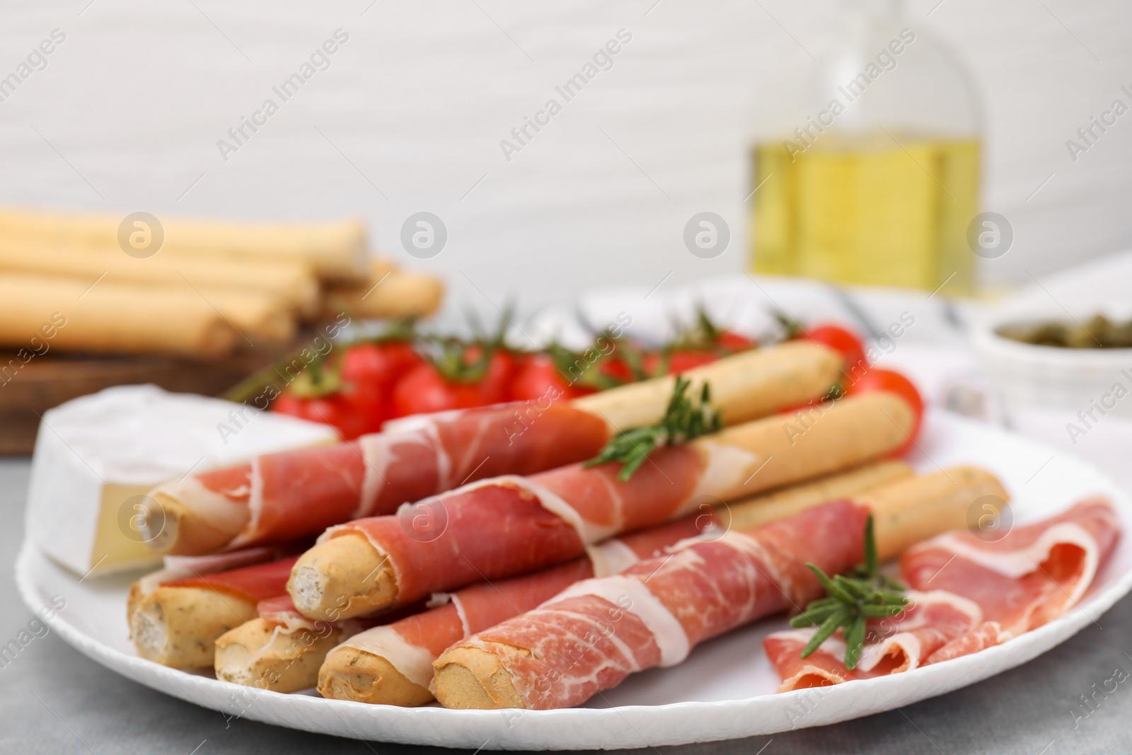 Photo of Plate of delicious grissini sticks with prosciutto, cheese and tomatoes on light grey table, closeup